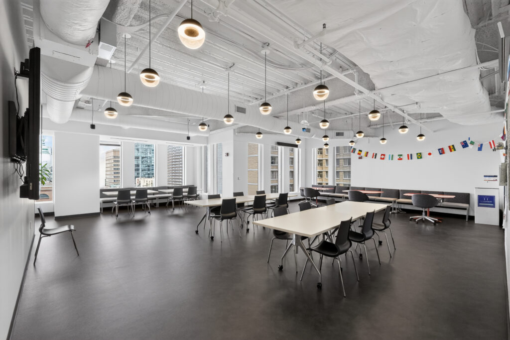 A modern office breakroom with white ceilings, pendant lighting, rows of tables and chairs, and a line of international flags on the back wall.