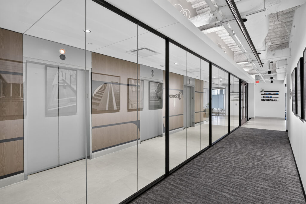 Office elevator lobby enclosed by glass walls with framed architectural photography on the wood-paneled elevator doors.