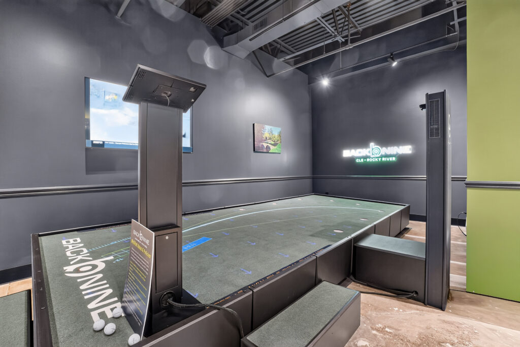 Indoor golf simulator with a putting green, two ball dispensers, and a Back Nine brand sign on the wall.