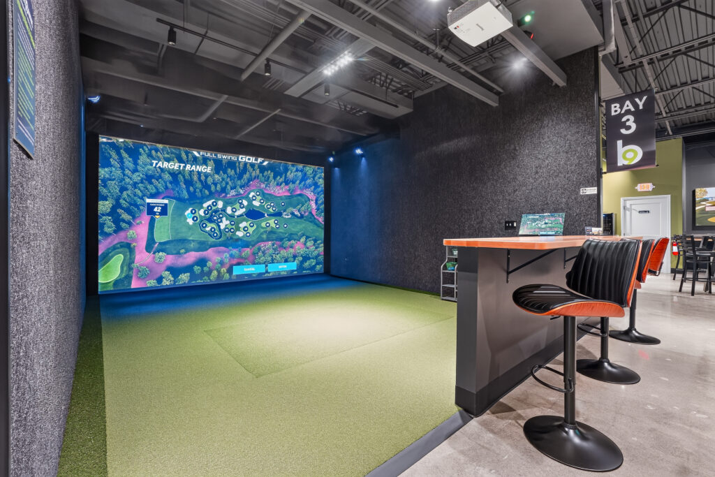Indoor golf simulator bay featuring a large screen displaying a target range map, a high-top counter with two stools, and a sign labeled “Bay 3.”