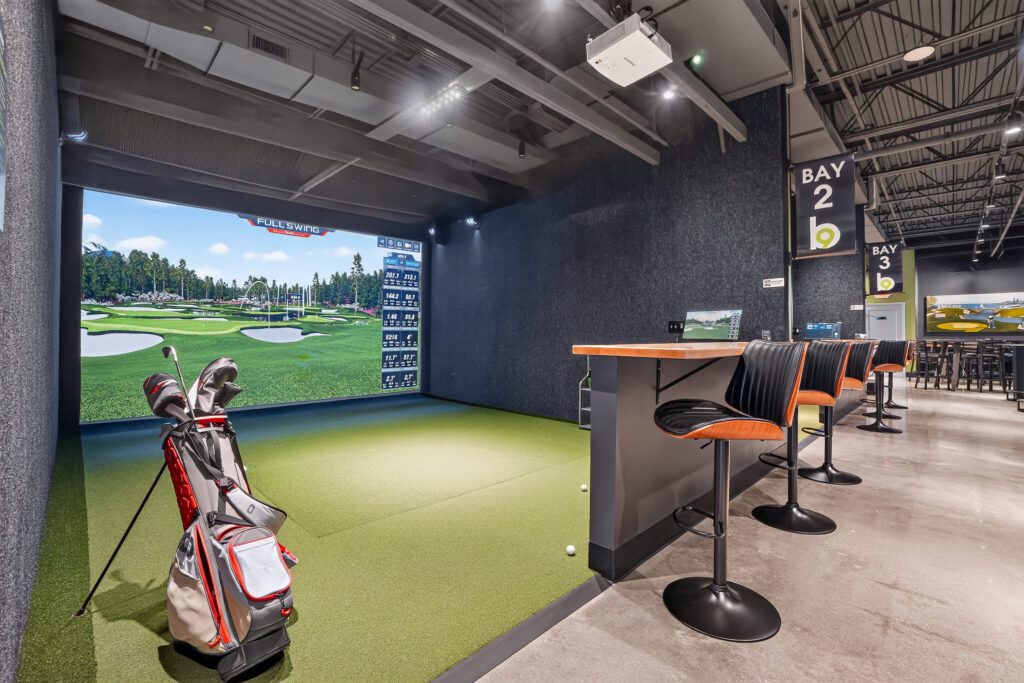 An indoor golf simulator bay with a screen displaying a course, a golf bag in the foreground, and a high-top counter with bar stools.
