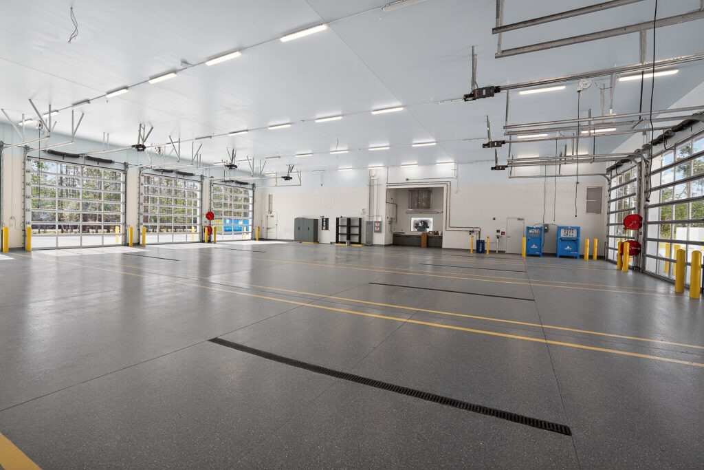 Large, empty vehicle maintenance garage with gray concrete floors, overhead glass doors, and yellow safety bollards.