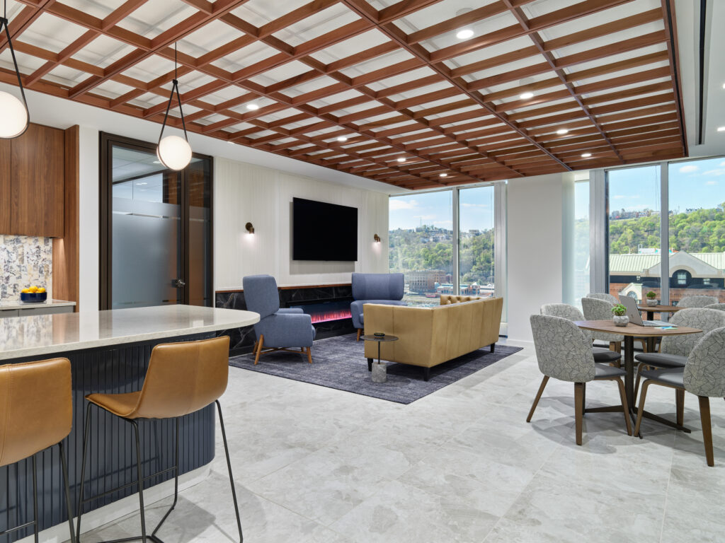 Modern office lounge featuring a kitchen island, blue armchairs, a tan sofa facing a fireplace and television, and a small round dining table.