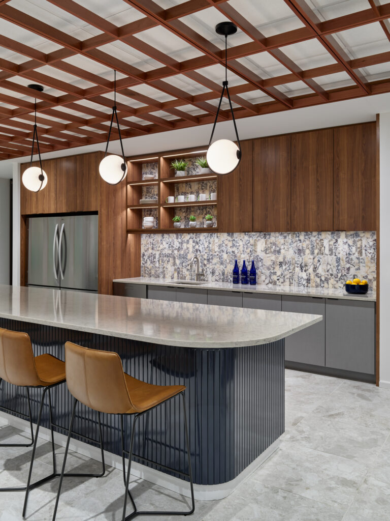 Modern kitchen with a dark blue island, leather bar stools, walnut cabinetry, and a stone-tiled backsplash.