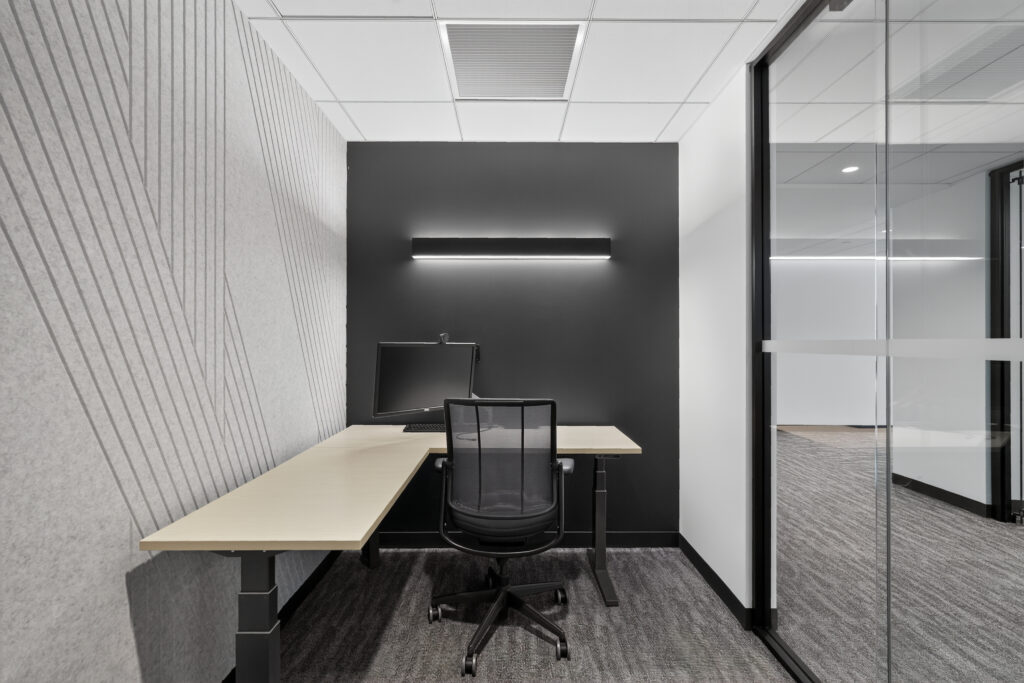 A small office cubicle with a desk, computer monitor, and ergonomic chair against a black wall with a horizontal light fixture.