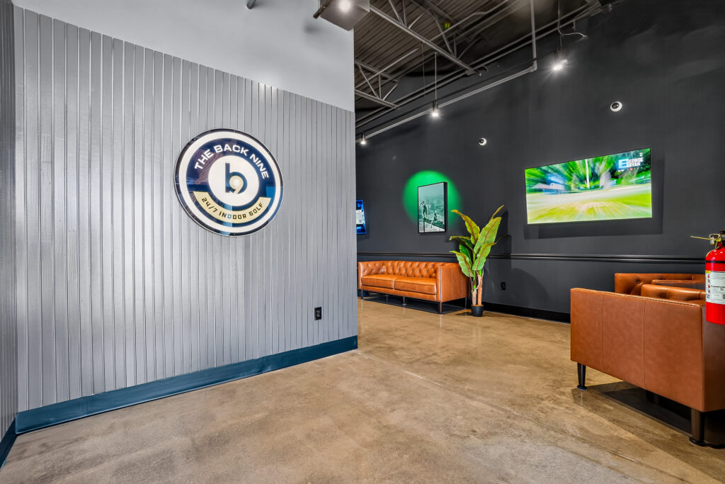 Interior of The Back Nine indoor golf facility featuring a lounge area with leather sofas, a mounted television, and the company logo on a gray paneled wall.