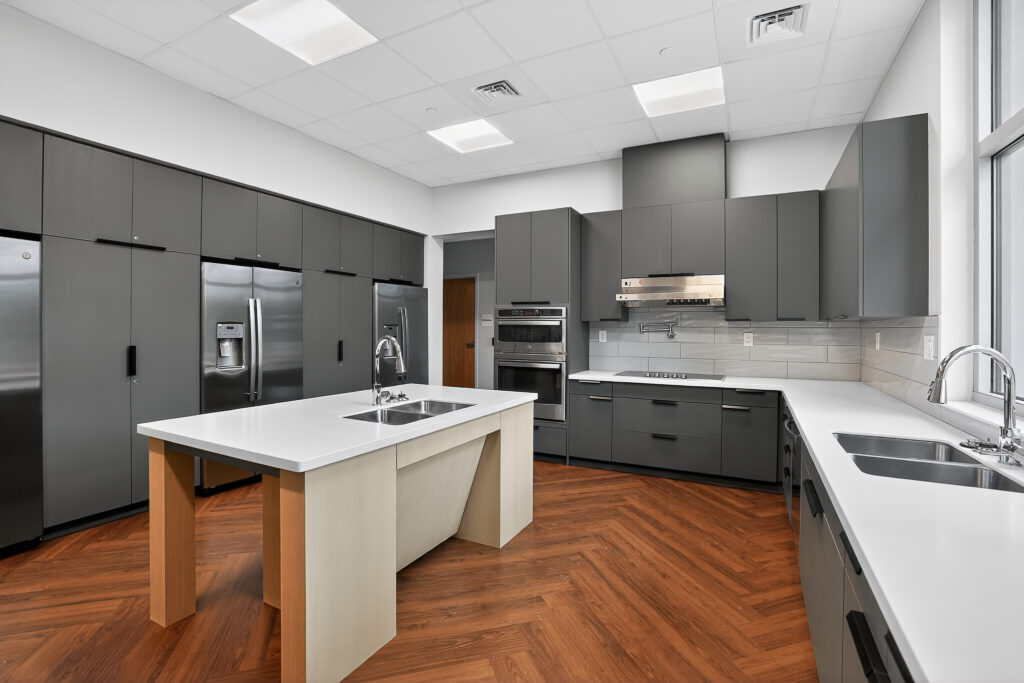 Modern kitchen with charcoal gray cabinetry, white countertops, two sinks, stainless steel appliances, and wood-patterned flooring.