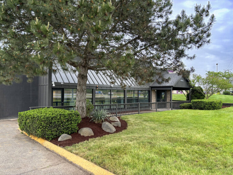 A modern, dark-sided building with the number 914 above the front door, situated behind a tree and a small garden bed.