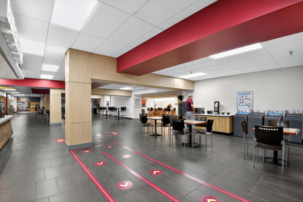 A cafeteria space with tables and chairs, featuring floor markings with directional arrows leading toward a service counter.