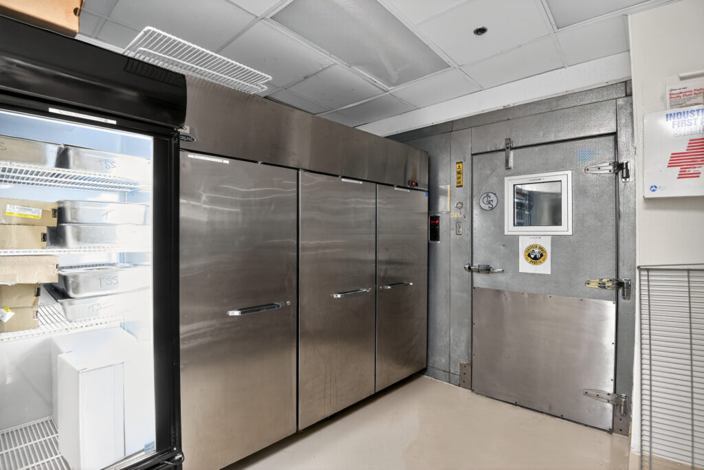 Commercial kitchen with a glass-front refrigerator, stainless steel reach-in coolers, and a walk-in freezer door with a first aid kit on the wall.