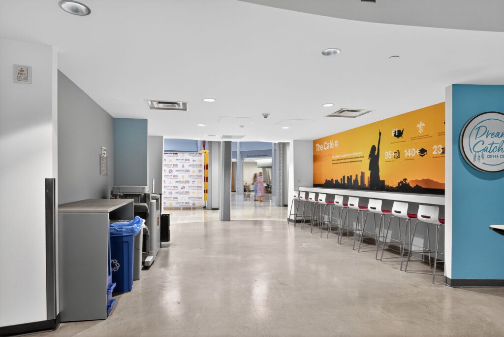 A cafe seating area with a row of stools against a wall mural, next to a hallway and a recycling station.