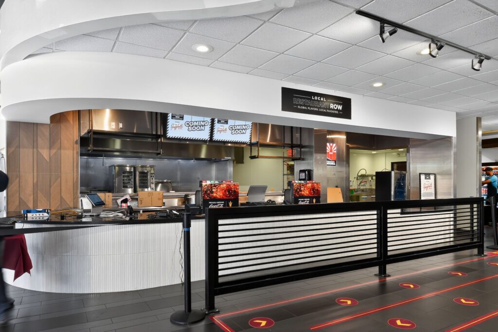 A commercial kitchen service counter labeled “Local Restaurant Row,” featuring digital menu boards that display “Coming Soon.”
