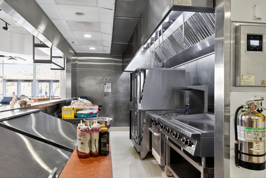 A professional kitchen featuring a commercial oven and flat-top grill, with condiment bottles on a counter and a fire extinguisher mounted on the wall.