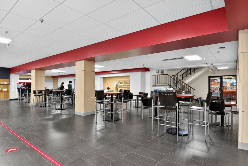 An interior view of a university dining area with high-top tables, stools, a red accent wall, and a service desk labeled “Sun Devil ID Services.”