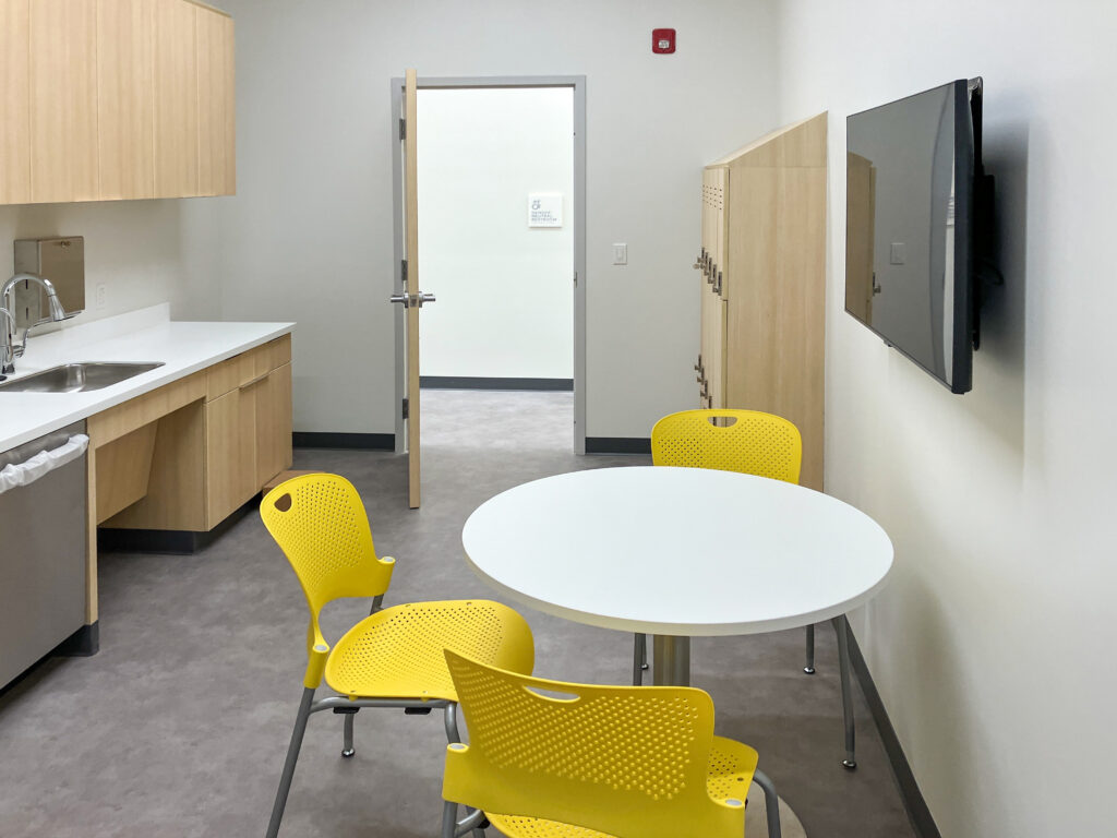 A breakroom with a round white table, three yellow chairs, a kitchenette with a sink, wooden lockers, and a wall-mounted television.