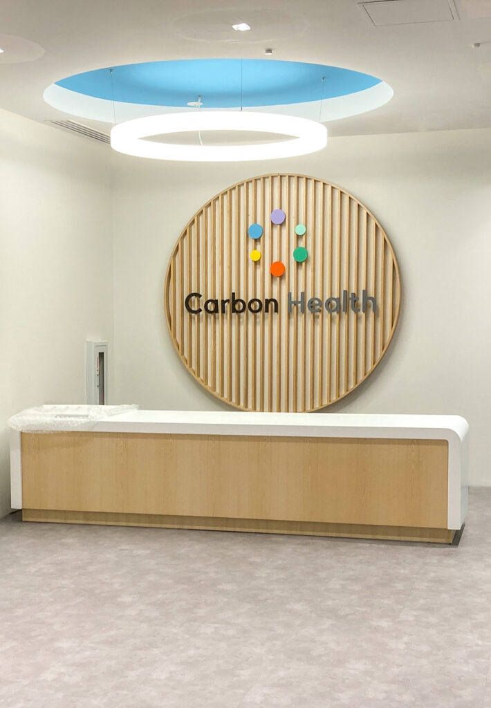 Carbon Health reception desk with a circular wood-slat logo display on the wall behind it.