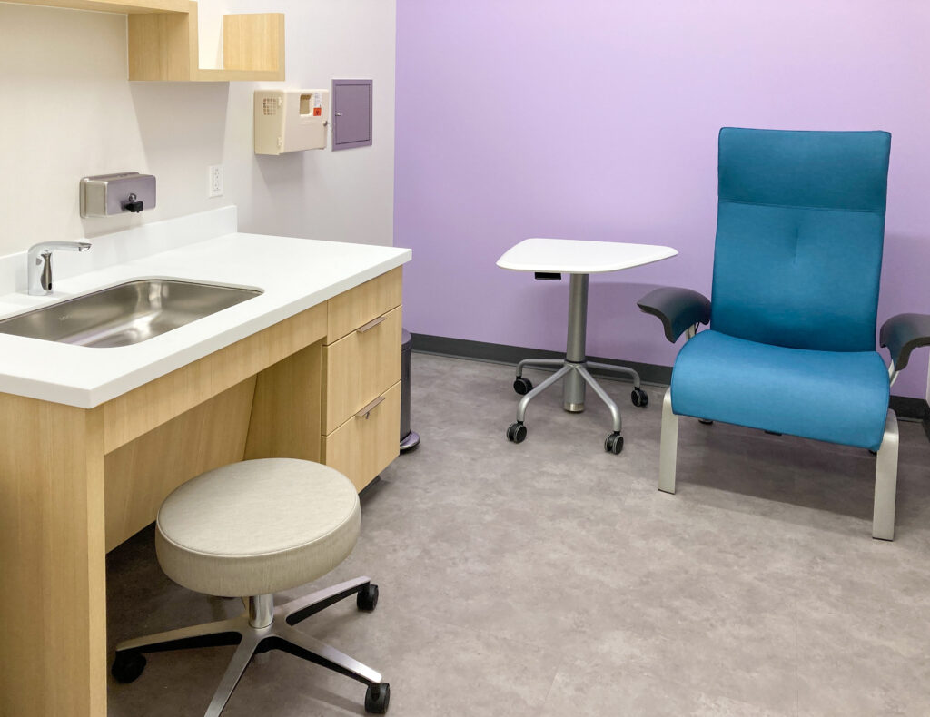 A medical exam room featuring a sink, a beige rolling stool, a small rolling table, and a blue clinical chair against a light purple wall.