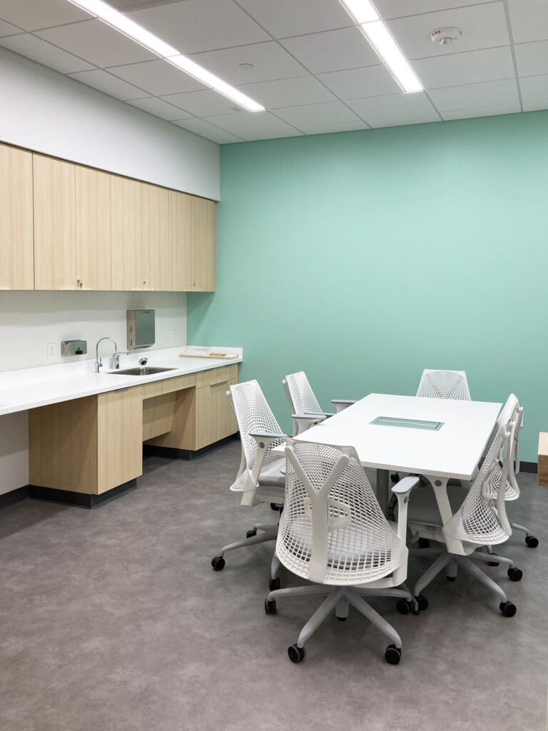 A meeting room with a white table and six modern white chairs, set against a teal wall next to a kitchen cabinet and sink area.