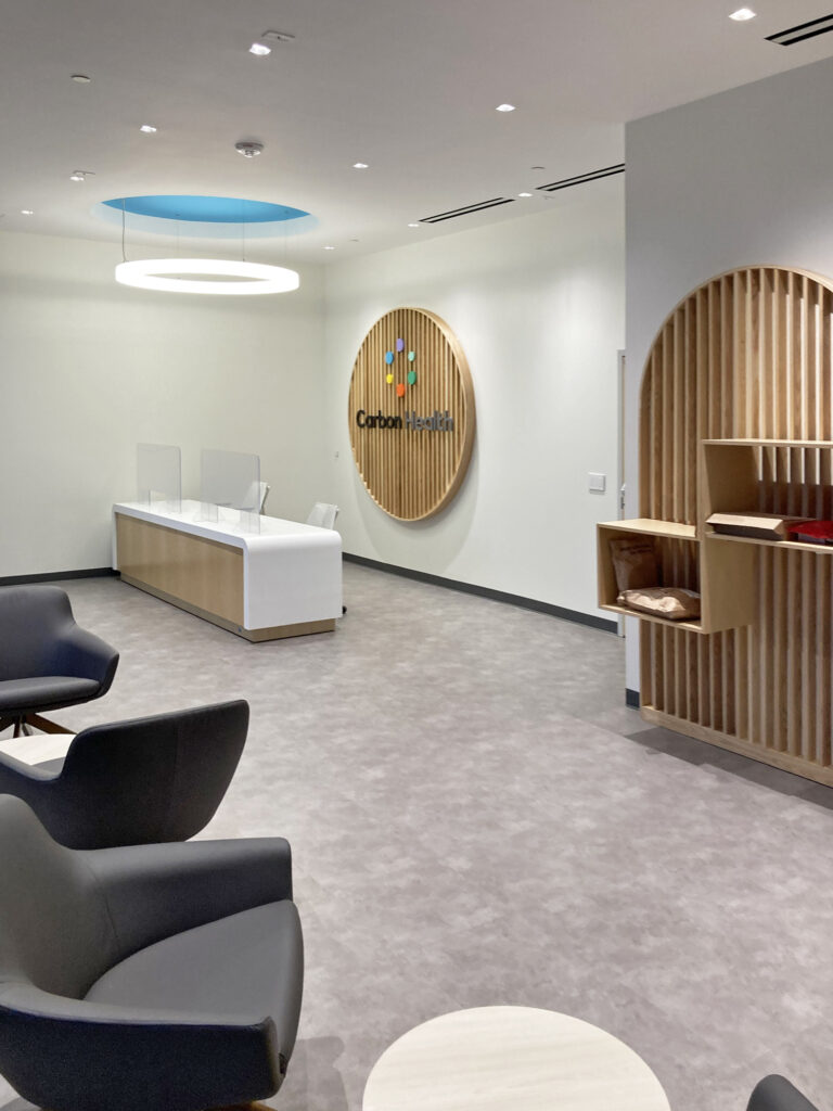 A Carbon Health reception area with a white front desk, gray chairs, and a circular wooden sign with the company logo on the wall.