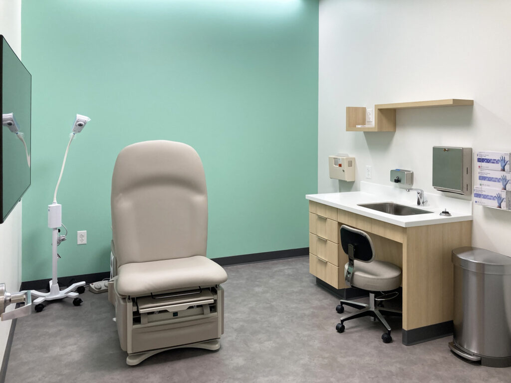 A medical exam room featuring a beige exam table, a desk with a sink, a stool, and a mobile examination light.
