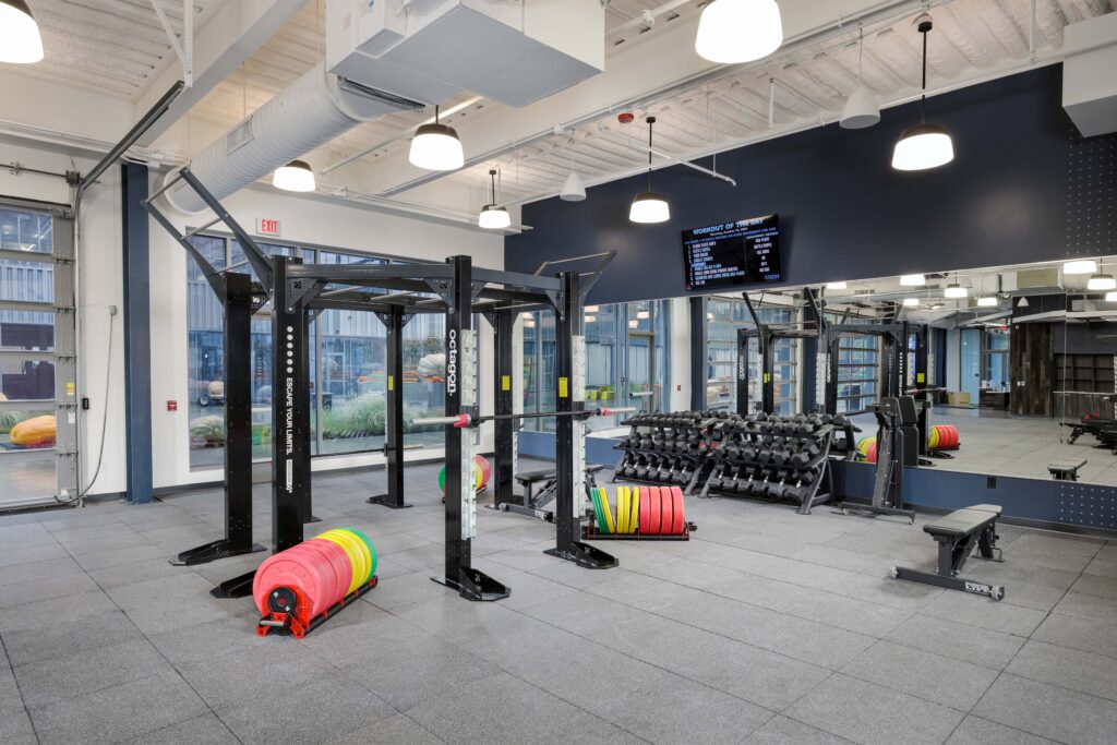 A gym interior featuring an Octagon fitness rig, racks of dumbbells, weight plates on the floor, and a wall-mounted screen displaying a workout plan.