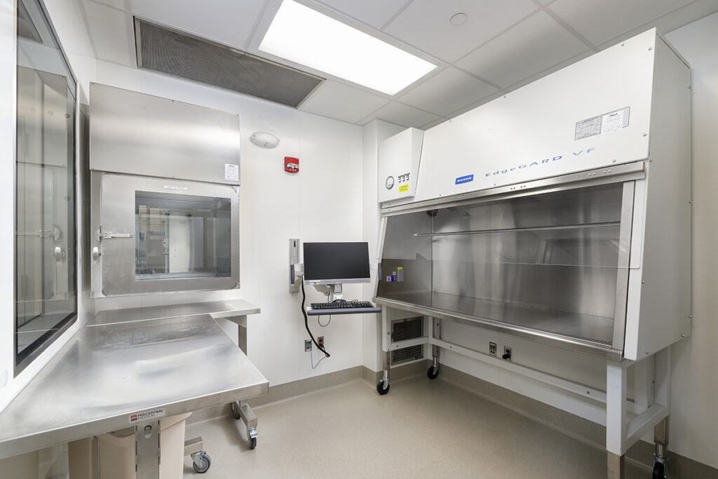 Laboratory room with a stainless steel pass-through chamber, a mounted computer monitor and keyboard, and a large Baker EdgeGARD laminar flow hood.