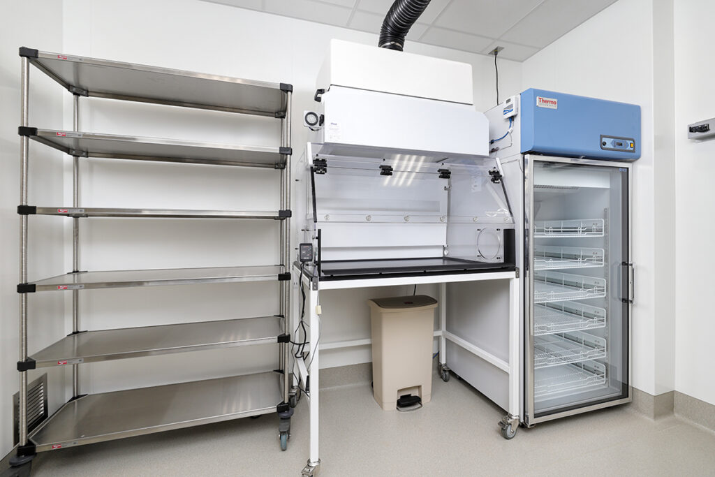 A laboratory workspace featuring a tall metal shelving unit, a white fume hood with a trash bin underneath, and a Thermo Scientific laboratory refrigerator with a glass door.