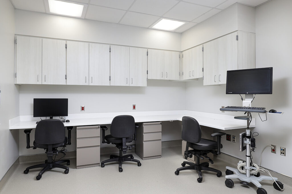 An office workstation with a wrap-around desk, three rolling chairs, upper cabinets, and a computer on a mobile stand.