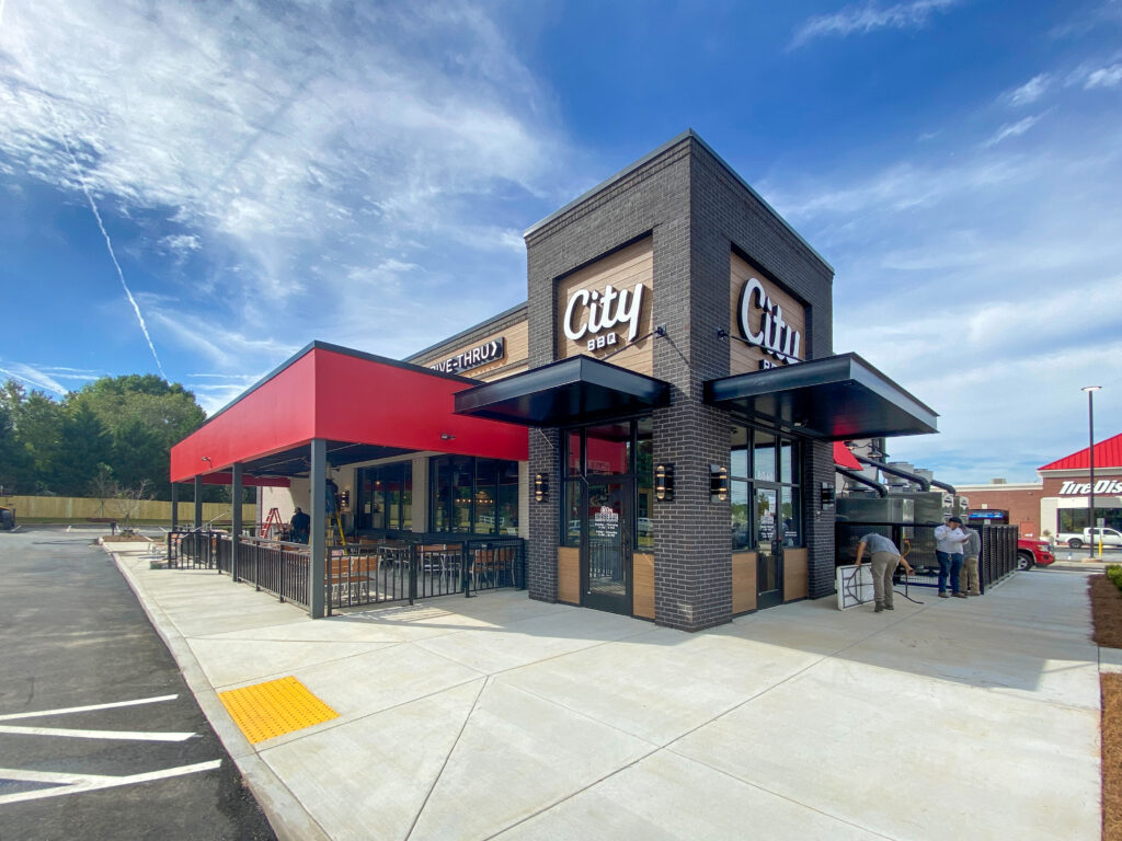 Exterior view of a City BBQ restaurant building with an outdoor patio and two workers installing a sign.