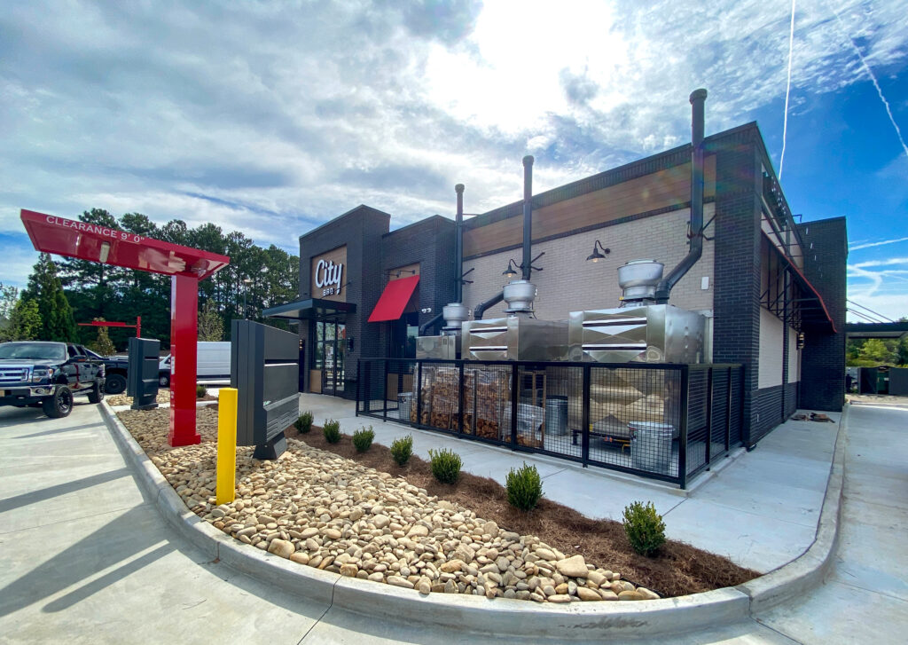 Exterior of a City BBQ restaurant showing the drive-thru lane and large outdoor smokers behind a metal fence.