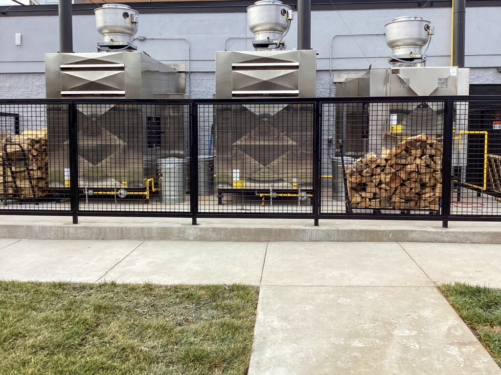 Three industrial kitchen exhaust vents installed against a building, with stacks of firewood stored behind a black metal fence.
