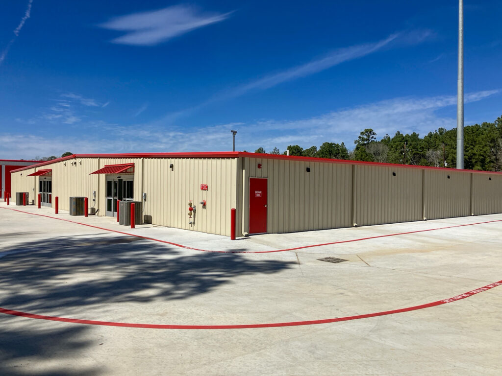 Tan commercial storage building with red trim and entry doors, situated in a concrete parking lot under a blue sky.