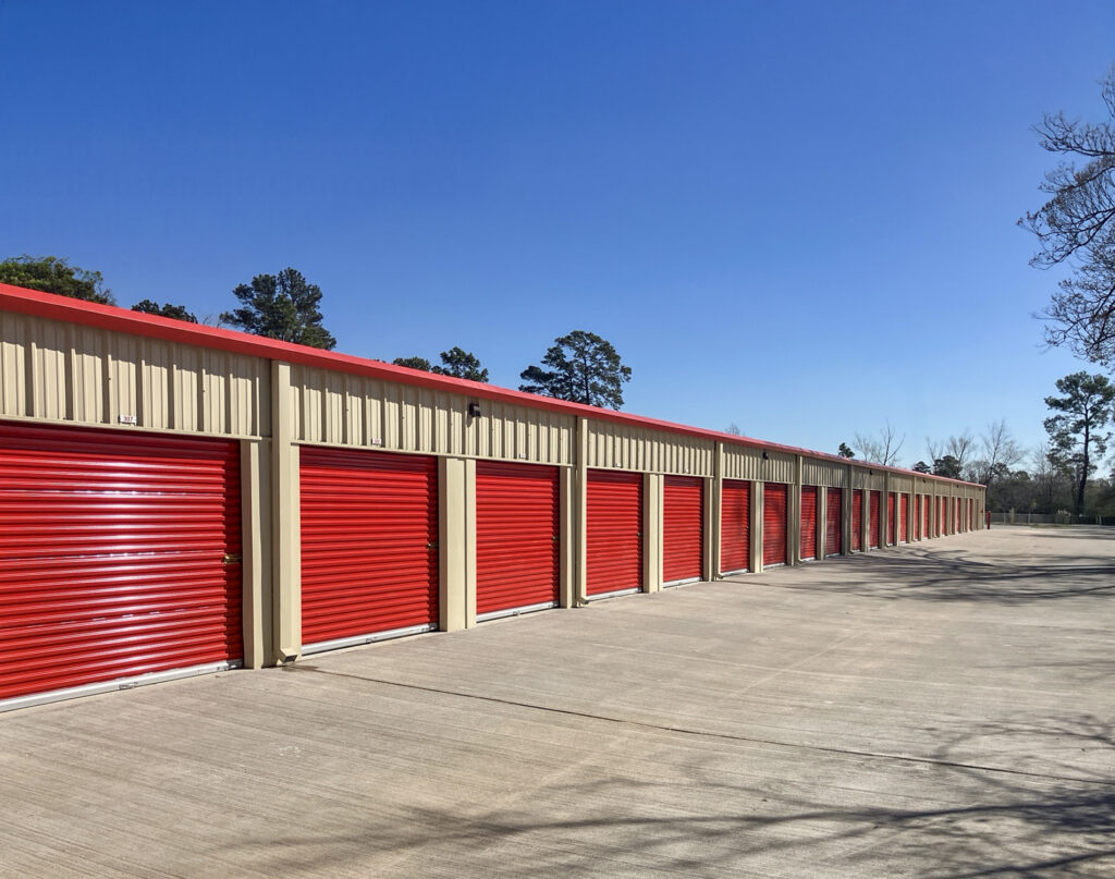 A long row of outdoor self-storage units with red roll-up doors and beige metal siding under a clear blue sky.