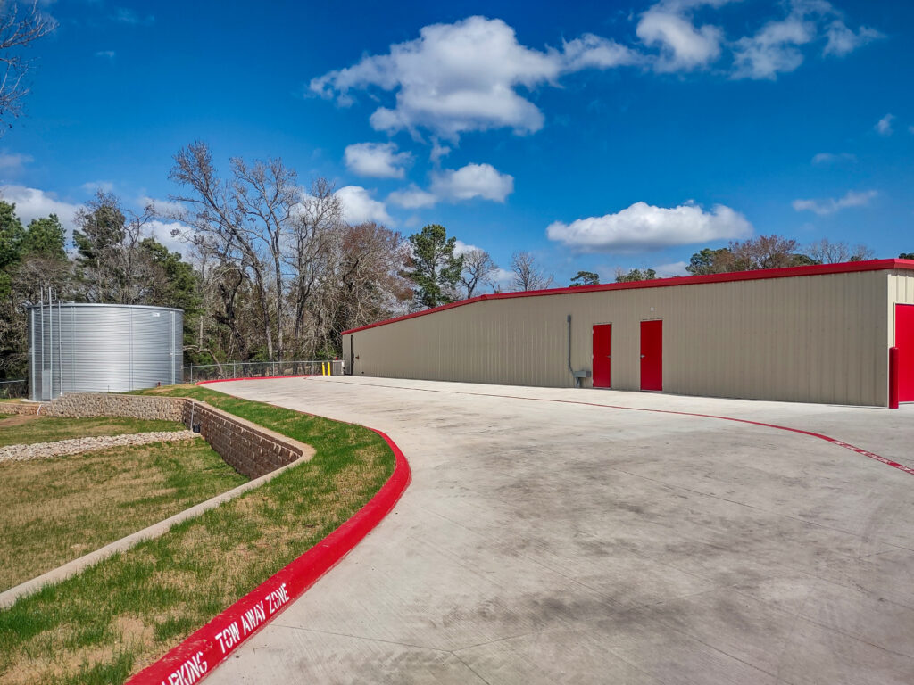 A tan metal building with red doors next to a concrete driveway and a large cylindrical water tank.