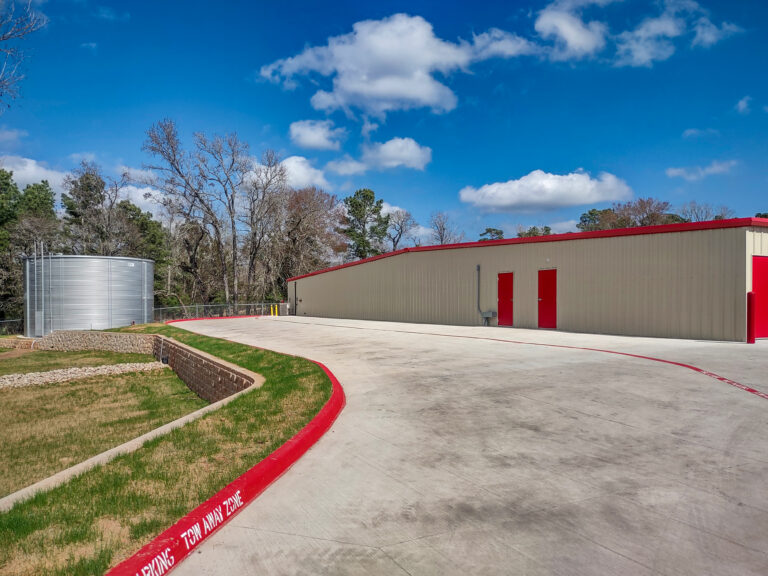 A tan metal building with red doors next to a concrete driveway and a large cylindrical water tank.