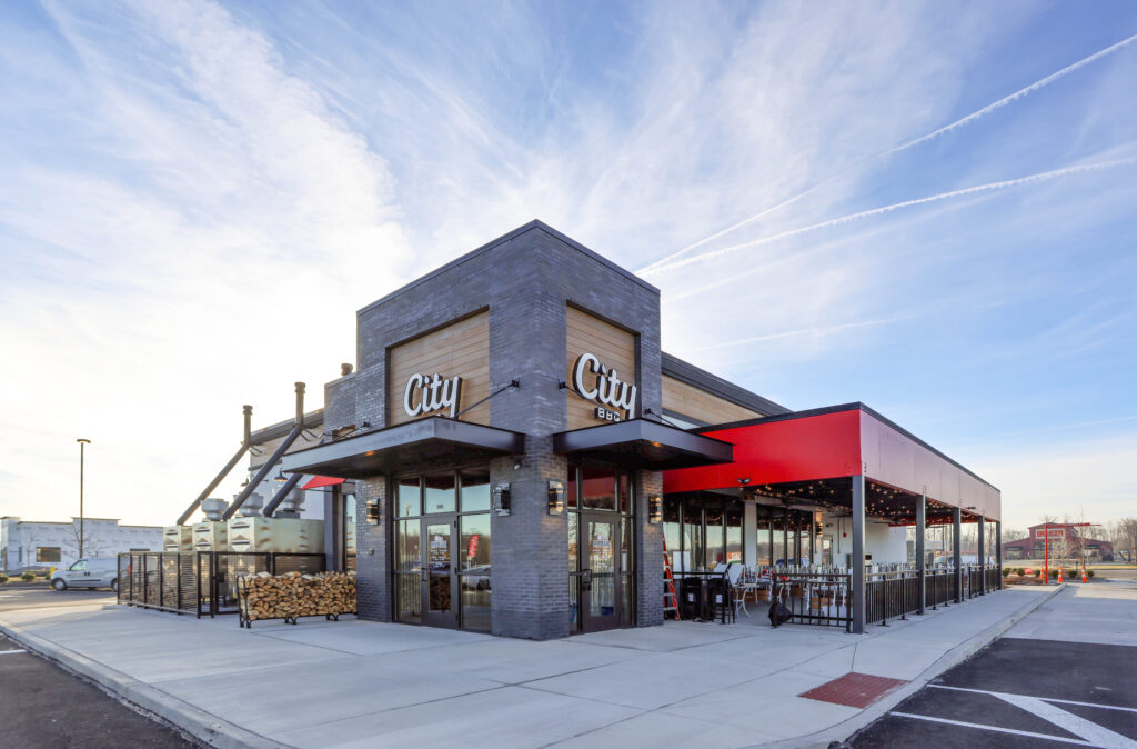 Exterior of a City BBQ restaurant with a grey brick corner entrance and a red-roofed outdoor patio.