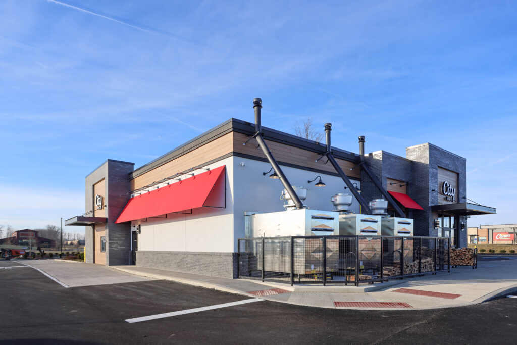 Exterior view of a City Barbeque restaurant building with large metal ventilation units and stacks of firewood enclosed by a fence.