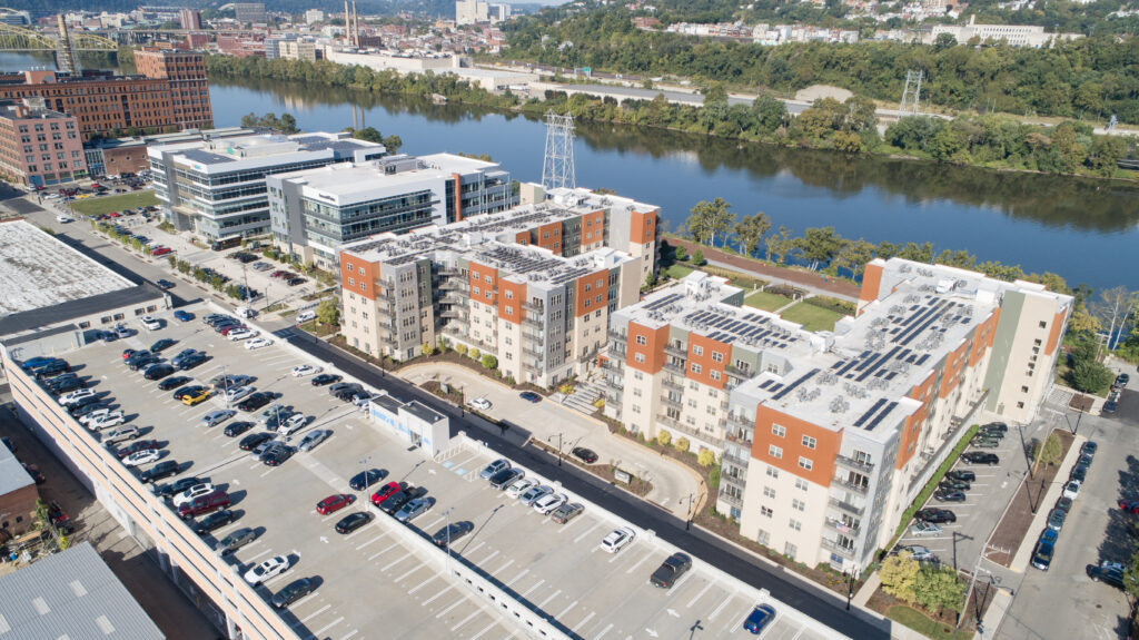 Aerial view of an apartment complex with solar panels on the roof, adjacent to a parking lot and a river in a city.