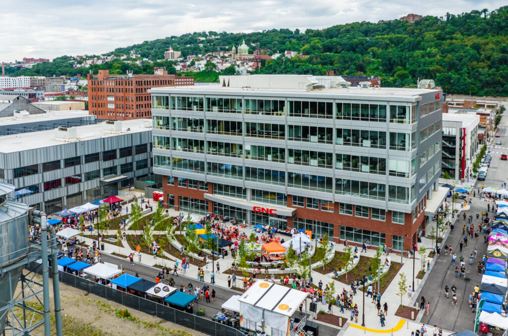 A GNC corporate building surrounded by an outdoor event with vendor tents and a crowd of people.