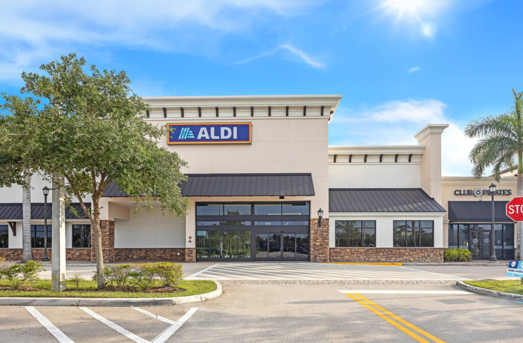The exterior of an Aldi grocery store with a tan facade and dark metal awnings under a blue sky.