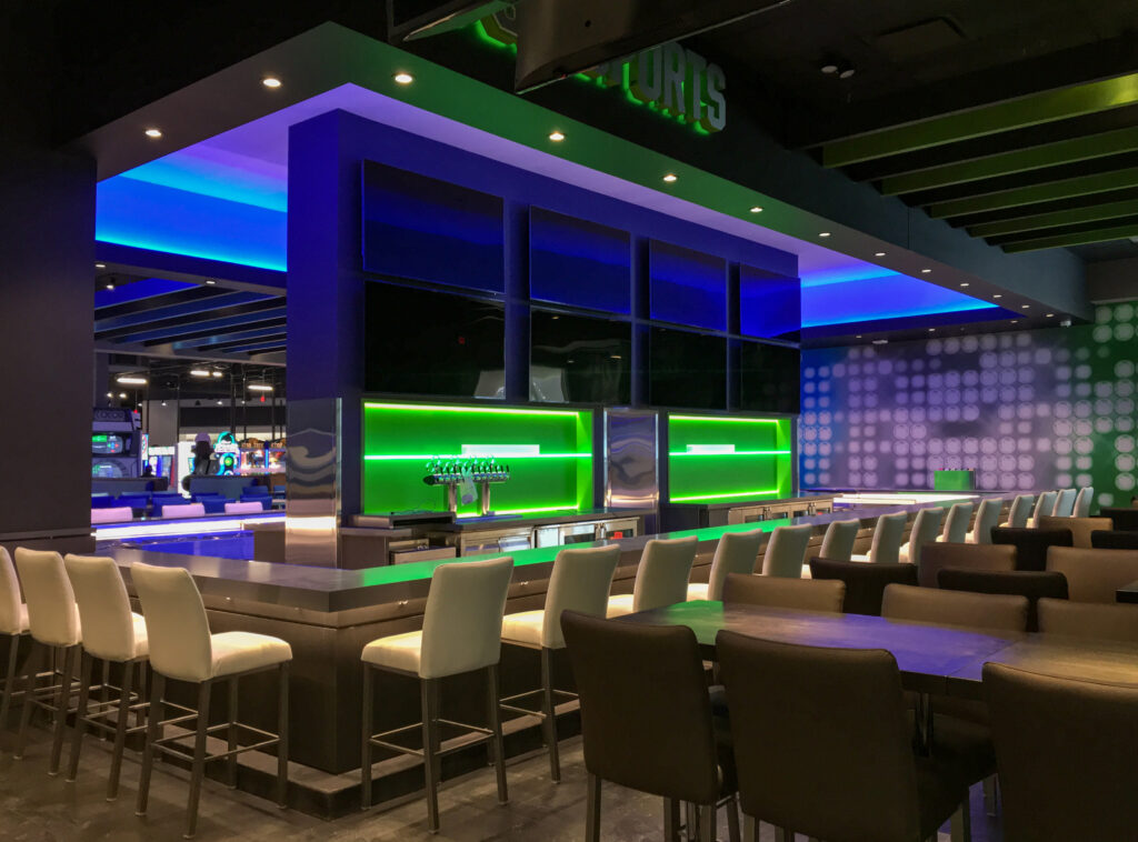 A modern sports bar with white bar stools, a backlit green counter, and multiple television screens mounted on a dark blue wall.