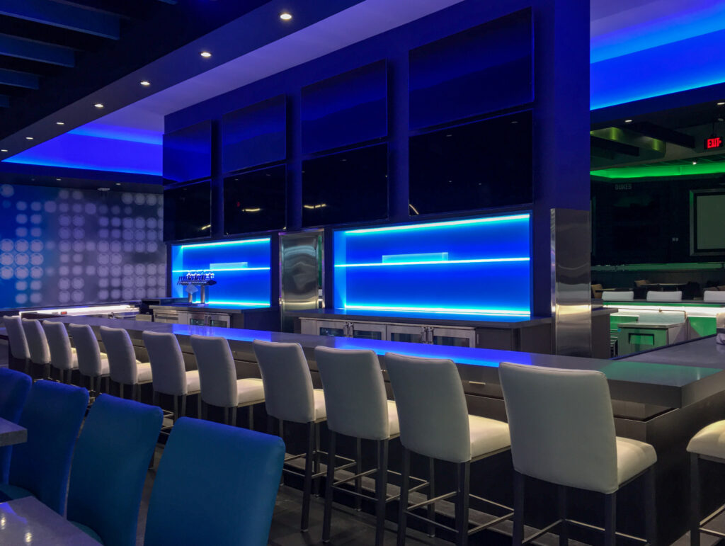 A modern bar with white stools facing a counter lit by blue neon, with multiple dark television screens mounted on the wall above.