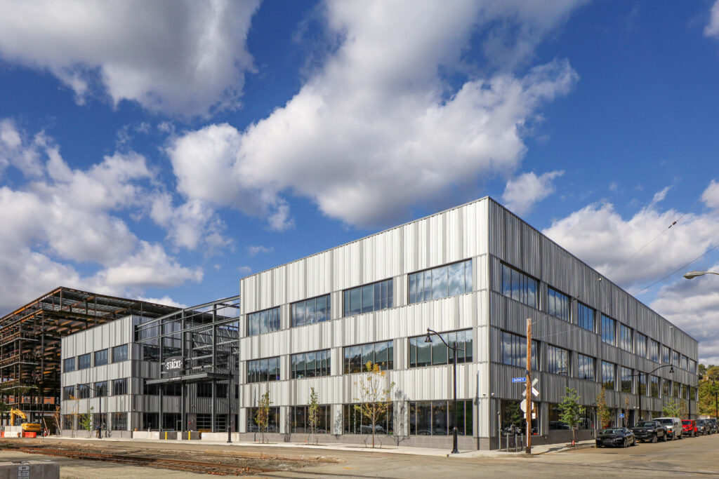 A three-story modern building with a metal facade, featuring a partially constructed steel frame section labeled “THE STACKS.”