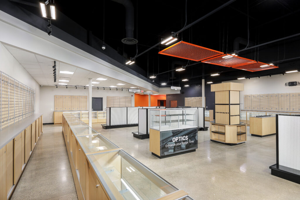 Interior of an empty retail store with glass display cases and shelving units, featuring a sign that reads “OPTICS Enhance your Range Time.”