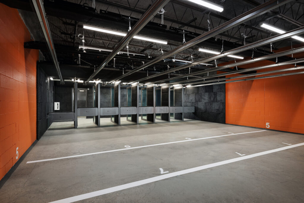 An indoor shooting range with multiple firing lanes, bright overhead lighting, and numbered markings on the concrete floor.