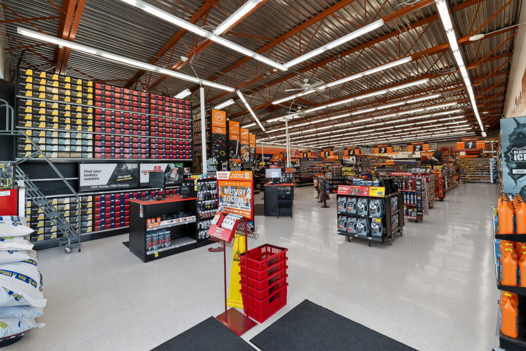 Interior view of an auto parts store with shelves of car batteries, motor oil, and a counter with a sign advertising a military discount.