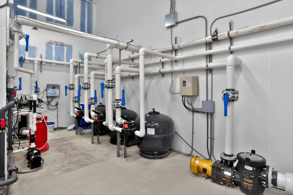 A mechanical room featuring a series of large black pool filters connected by white PVC piping, with several blue manual valves and electrical control boxes mounted on the wall.