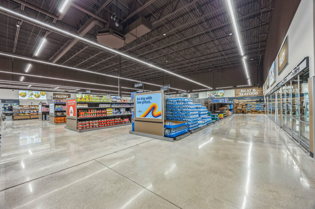 Interior view of a grocery store aisle featuring a sign that reads “Go big with low prices,” shelves of pantry goods, and a large display of bottled water.