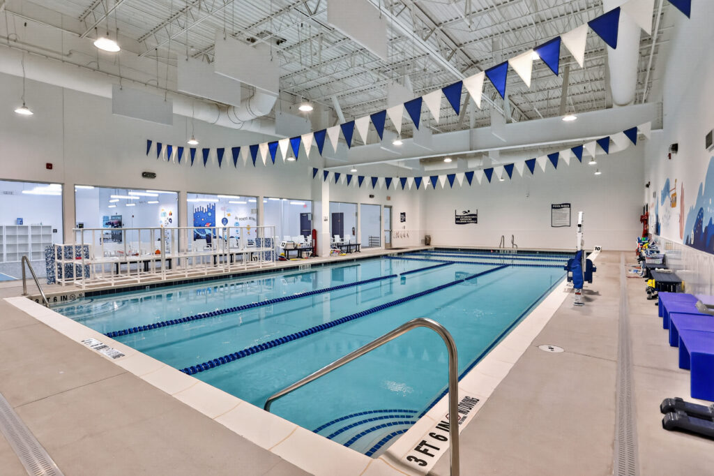 Indoor lap pool with blue lane markers, surrounded by a white deck, blue and white triangular pennants overhead, and glass walls looking into an adjacent room.