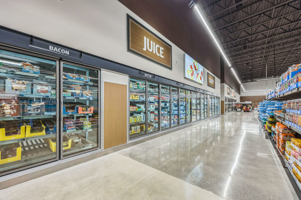 A grocery store aisle featuring long rows of refrigerated glass-door cabinets labeled for bacon, juice, butter, yogurt, and dairy.